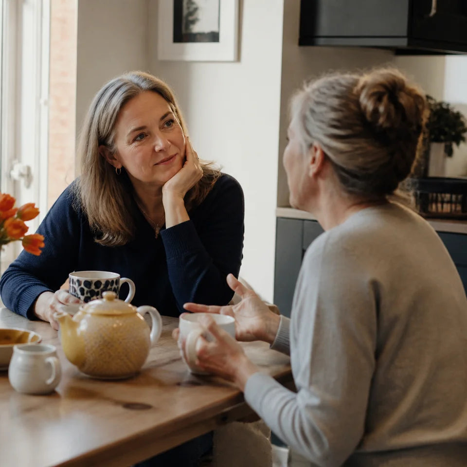 Two women in their fifties talking over tea at a kitchen table, seen from the side
